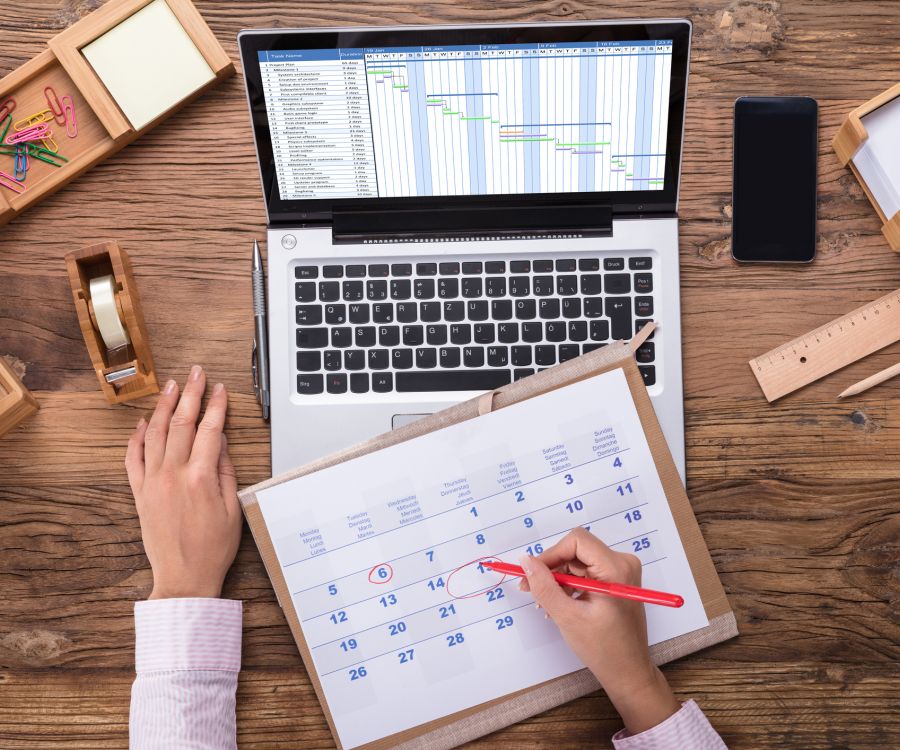 Person checking a physical calendar atop a laptop keyboard with project plan on screen.