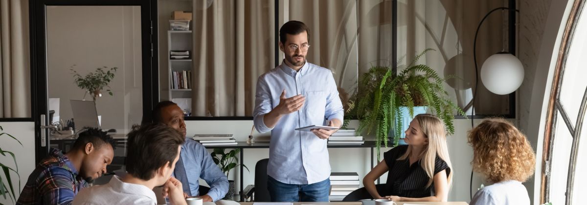 Meeting - man leading a team meeting with six people seated around a table in a modern office.